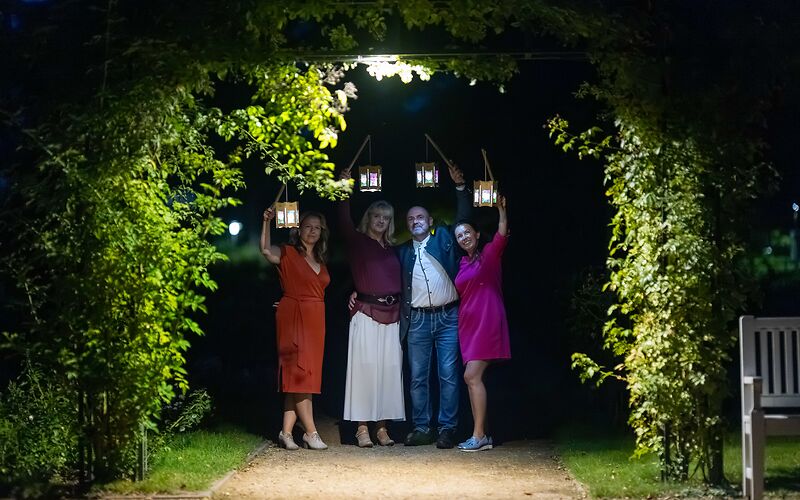 Eine Nachtaufnahme zeigt vier bunt gekleidete Personen mit leuchtenden Lampions unter einer beleuchteten Rosenpergola. Die stimmungsvolle Beleuchtung verleiht der Szene eine besondere Atmosphäre.