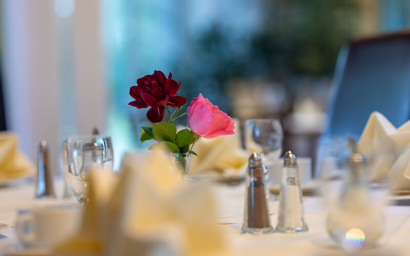 Ein stilvoll gedeckter Tisch im Restaurant Rosenflair. In der Mitte des Tisches stehen zwei Rosen in Rosa und Dunkelrot als dekorativer Blickfang.