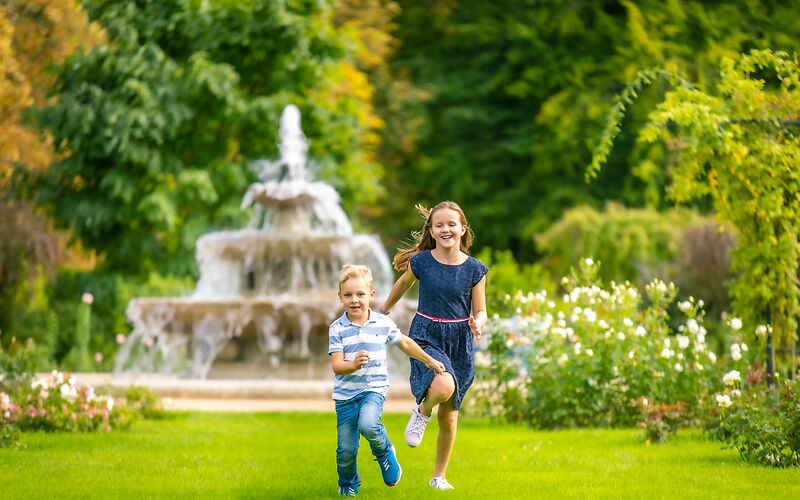 Das Foto zeigt ein Mädchen und einen Jungen, die lächelnd durch eine Wiese im Neurosengarten laufen. Im Hintergrund sind die Kaskaden mit fließendem Wasser sowie grün und orange gefärbte Bäume zu sehen.