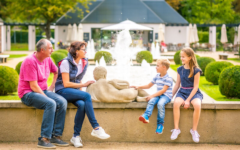 Das Foto zeigt Großeltern mit ihren Enkeln, die eine Pause vor den Großen Wasserspielen im Ostdeutschen Rosengarten genießen. Im Hintergrund sind ein Café und weitere Besuchende der Gartenanlage zu sehen. Die Szene vermittelt eine entspannte Atmosphäre inmitten der historischen Parklandschaft.