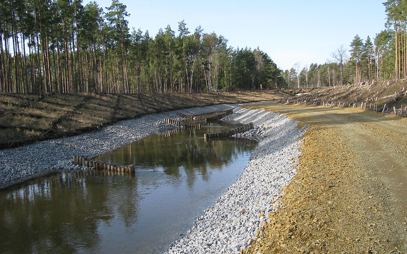 Wasser fließt durch einen Graben mit Boden aus vielen Steinen, rechts daneben Verläuft ein Sandweg. Links und rechts ist Wald.