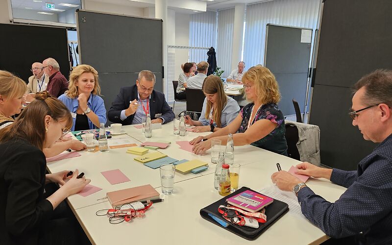 Frauen und Männer sitzen im Kreis am Tisch, vor ihnen bunte beschriebene Karten 