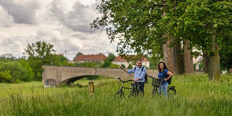 Eine Radreisende und ein Radreisender legen vor der historischen Langen Brücke in Forst (Lausitz) eine kurze Pause ein. Im Bild ist nur ein Teil der Brücke zu sehen. Im Hintergrund stehen große Bäume und die Häuser entlang des Kegeldamms.