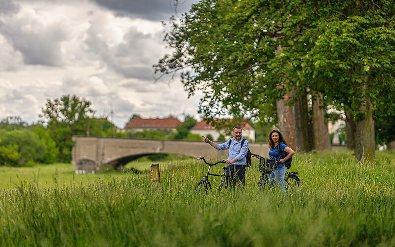 Eine Radreisende und ein Radreisender legen vor der historischen Langen Brücke in Forst (Lausitz) eine kurze Pause ein. Im Bild ist nur ein Teil der Brücke zu sehen. Im Hintergrund stehen große Bäume und die Häuser entlang des Kegeldamms.