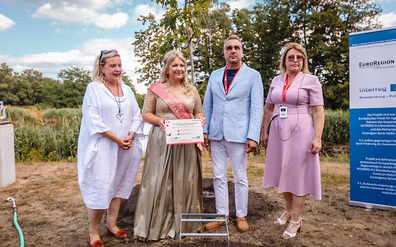 Rosenkönigin Stefanie III., Beata Kowalczuk, Janusz DudojÄ‡ und Simone Taubenek stehen vor einem der neu geplanzten Bäume. Stefanie die III. hält eine Infotafel zur Baumpflanzung in den Händen. Rechts steht ein Banner zum Fördermittelprojekt Interreg.