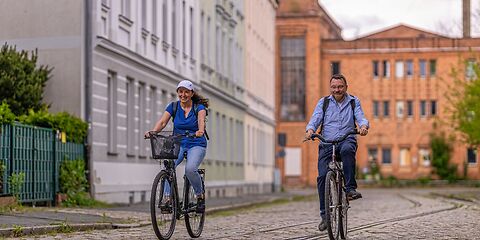 Ein Mann und eine Frau fahren auf einer gepflasterten Straße nebeneinander Fahrrad. Auf der Straße sind Schienen verlegt. Links steht ein Wohnhaus und im Hintergrund ist eine altes Fabrikgebäude aus Backstein zu sehen.