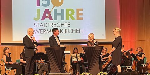 zu sehen ist eine Bühne, auf der eine Podiumsdiskussion abgehalten wird, Bürgermeisterin Simone Taubenek hat das Mikrofon