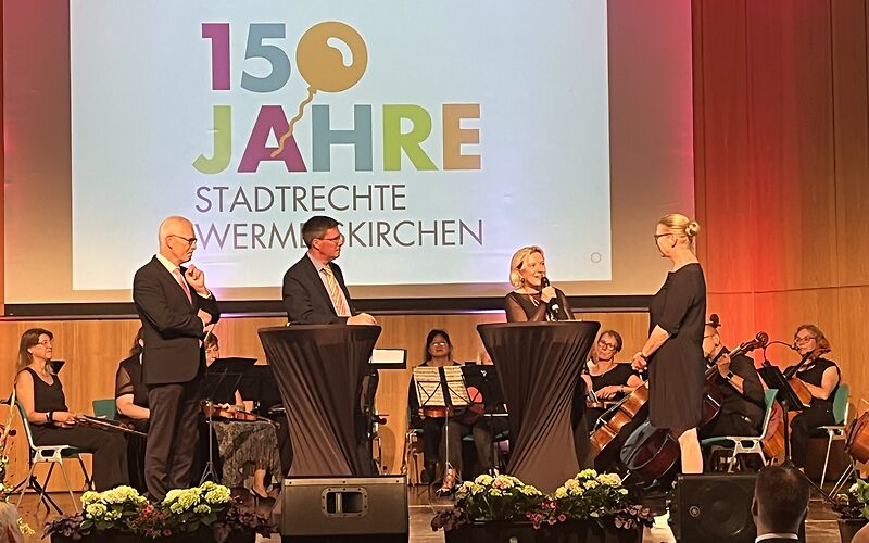 zu sehen ist eine Bühne, auf der eine Podiumsdiskussion abgehalten wird, Bürgermeisterin Simone Taubenek hat das Mikrofon