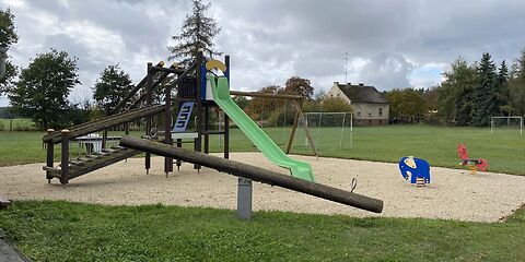 Auf einer Sandfläche stehen ein Klettergerüst aus Holz, an dem eine grüne Rutsche befestigt ist und zwei Federwippen (rotes Pferd und blauer Elefant). Davor steht auf der Wiesen eine Holzwippe.