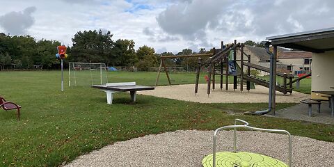 Vorn ist ein Karussel (Drehscheibe mit drei zur Mitte gebogenen Metallstangen) eine Tischtennisplatte, ein Klettergerüst und ein Fußballtor.