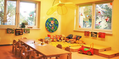 Zu sehen ist der Gruppenraum der Ameisengruppe, im Vordergrund stehen Tische mit Stühlen rundherum, in der Ecke liegen Matratzen mit Kissen und Kuscheltieren, das Zimmer ist hell und freundlich in gelb gehalten.
