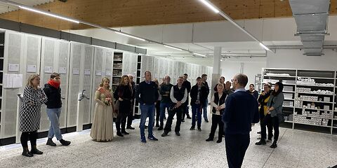 Blick in das Magazin: Schiebbare Regale mit Archivgut und offene Lagerregale. Im Raum steht die Gruppe Besucher.