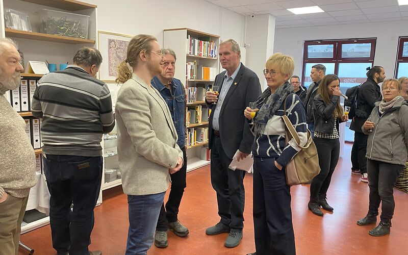 Im Lesesaal stehen die Besucher der Archiveröffnung. An der Wand links befinden sich Regale mit Büchern/Ordnern.