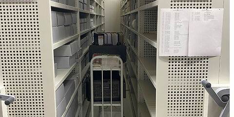 Blick zwischen zwei Regale mit Archivgut, in der Mitte steht ein Wagen mit Ordnern. An den Seiten der Schränke sind Kurbeln befestigt, um sie verschieben zu können.