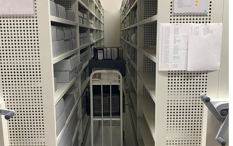 Blick zwischen zwei Regale mit Archivgut, in der Mitte steht ein Wagen mit Ordnern. An den Seiten der Schränke sind Kurbeln befestigt, um sie verschieben zu können.