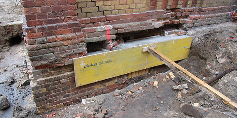 Ausbesserungen Öffnungen: ein Brett wird mit einer Stütze an die Mauer gedrückt. Der hinter den Ziegeln  entstandende Raum wird mit Beton gefüllt