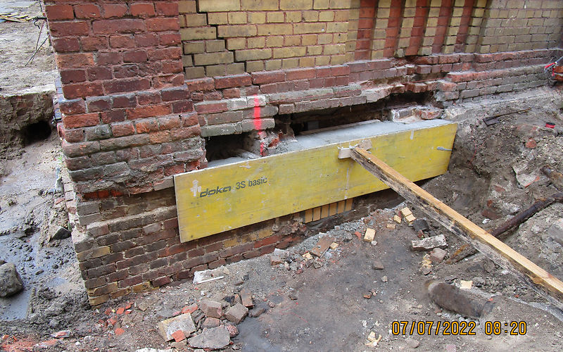 Ausbesserungen Öffnungen: ein Brett wird mit einer Stütze an die Mauer gedrückt. Der hinter den Ziegeln  entstandende Raum wird mit Beton gefüllt
