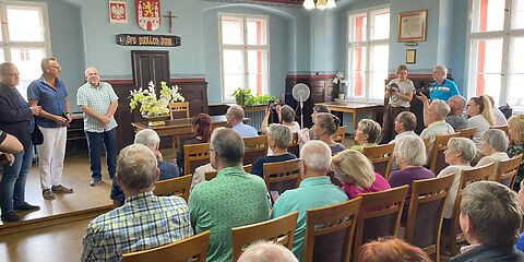 In einem Saal sind mehrere Stuhlreihen aufgebaut, auf denen Personen sitzen. Der hinteren Teil des Raumes ist leicht erhöt (eine Stufe) und auf diesem Teil steht ein Tisch mit einem leeren Stuhl dahinter. Mittig an der Wand hängt das Wappen der Stadt Lubsko.