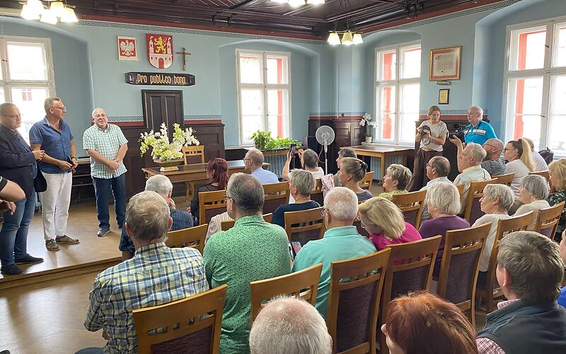In einem Saal sind mehrere Stuhlreihen aufgebaut, auf denen Personen sitzen. Der hinteren Teil des Raumes ist leicht erhöt (eine Stufe) und auf diesem Teil steht ein Tisch mit einem leeren Stuhl dahinter. Mittig an der Wand hängt das Wappen der Stadt Lubsko.