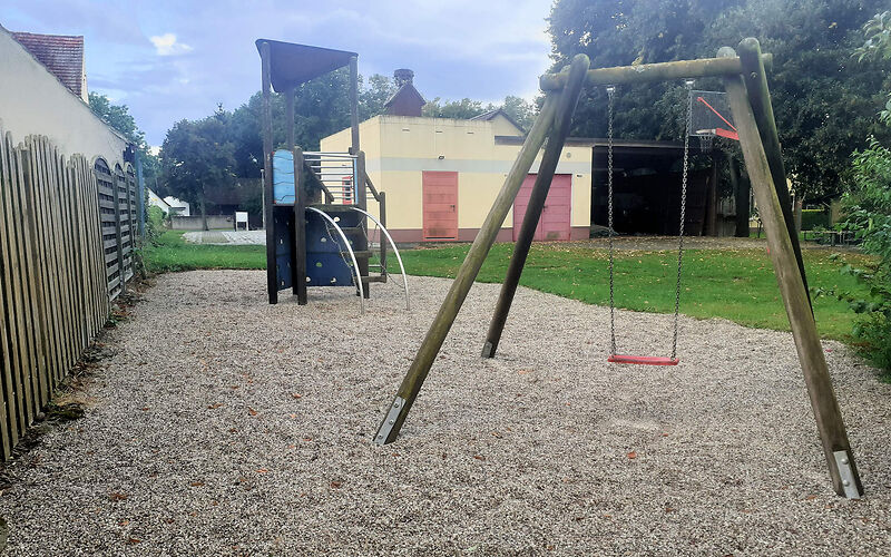 Auf einem Platz mit feinem Kies steht rechts eine an einem Holsgerüst befestigte Schaukel und links ein Klettergerüst mit einem Aufstieg aus 3 an gebogenden Rohren befestigten Holzstufen.