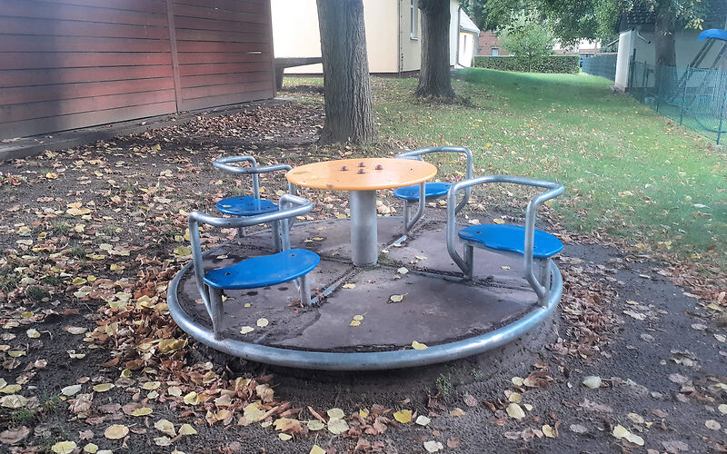 Ein rundes Karussel mit 4 blauen sitzen und einer Drehscheibe in der Mitte. Drum herum liegen bunte Blätter.