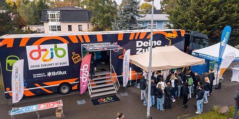, Foto:  Studio 2.0 - Christian Swiekatowski Stand auf dem Tag des offenen Unternehmens: Ein LKW, der durch eine Treppe mittig begehbar ist mit der Aufschrift "deine Zukunft ist bunt", davor stehen 2 Pavillione. Unter dem vorderen Pavillion steht eine große Gruppe Jugendlicher.
