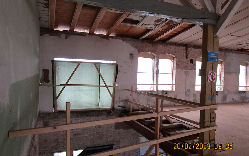 Auf der linken Seite im Hauptgebäude ist eine Deckenöffnung für den Lastenaufzug und auf der rechten Seite sind Fenster zu sehen. Ein großer Holzträger stützt die darüber liegende Decke ab. Die Deckenöffnung ist mit einer Holzumsperrung gesichert.