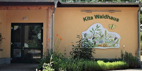 Zu sehen ist der Eingangsbereich der Kita, mit einer große zwei-flügelige Glastür, rechts davon ist der Schriftzug Kita Waldhaus aufgemalt und darunter ein großflächiges Bild mit Gräsern und einer Schnecke, darüber fliegt ein gelber Schmetterling