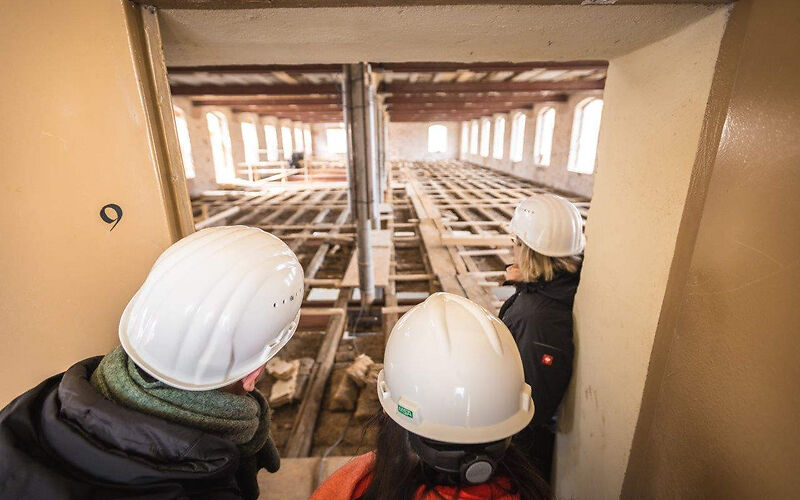 Blick in einen zu sanierenden Raum, zu sehen ist ein großer Raum im Rohbau, mit Industriefenstern, links und Rechts angeordnet, Fußbodenbeplankung und in einer Reihe angeordnete Träger für die Decke