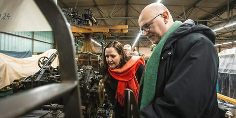 Zu sehen sind in einer Halle eingelagerte Textilmaschinen, Jörn Brunotte, Teamleiter des Museums erklärt Kulturministerin M. Schüle die Funktionsweise einer Textilmaschine