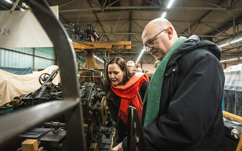 Zu sehen sind in einer Halle eingelagerte Textilmaschinen, Jörn Brunotte, Teamleiter des Museums erklärt Kulturministerin M. Schüle die Funktionsweise einer Textilmaschine