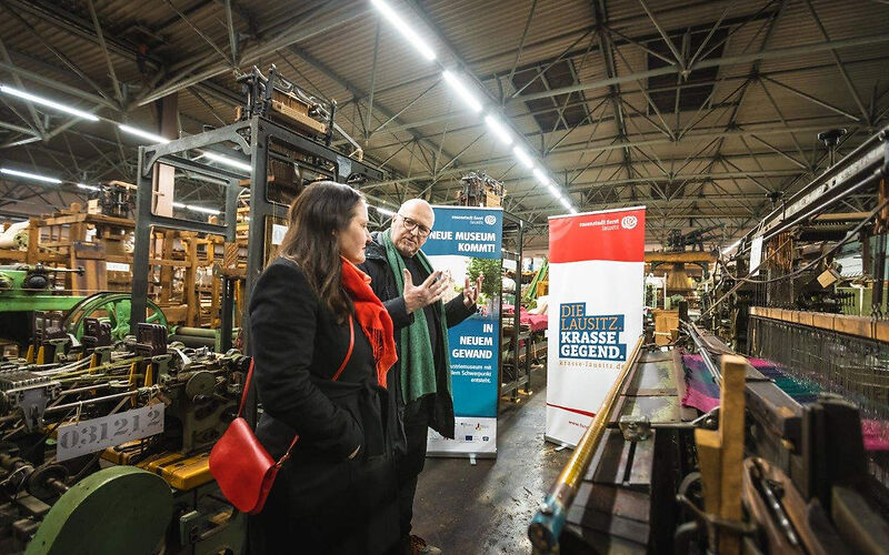 Kulturministerin des Landes Brandenburg M. Schüle im Gespräch mit Jörn Brunotte, Teamleiter des Brandenburgischen Textilmuseums, zu sehen sind in einer Halle eingelagerte Textilmaschinen aus dem Brandenburgischen Textilmuseums