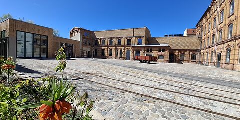 Innenhof des Museums (ein Backsteingebäudekomplex) mit Schienen auf gepflastertem Boden und Pflanzen im Vordergrund und einem Anhänger auf dem Hof