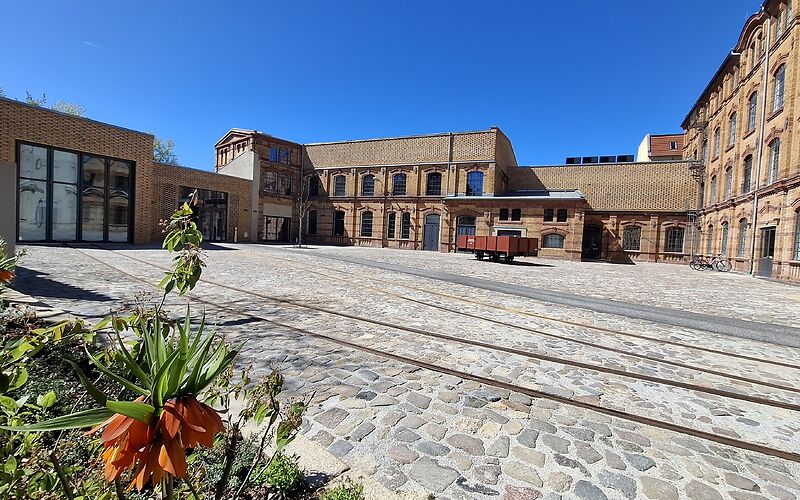 Innenhof des Museums (ein Backsteingebäudekomplex) mit Schienen auf gepflastertem Boden und Pflanzen im Vordergrund und einem Anhänger auf dem Hof
