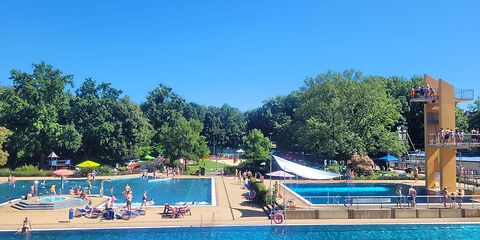 Blick über das Schwimmerbecken auf den Sprungturm mit dazugehörigem Becken und das Nichtschwimmerbecken, überall sind Kinder und Jugendliche. Die Sonne scheint.