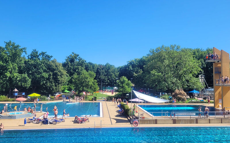 Blick über das Schwimmerbecken auf den Sprungturm mit dazugehörigem Becken und das Nichtschwimmerbecken, überall sind Kinder und Jugendliche. Die Sonne scheint.