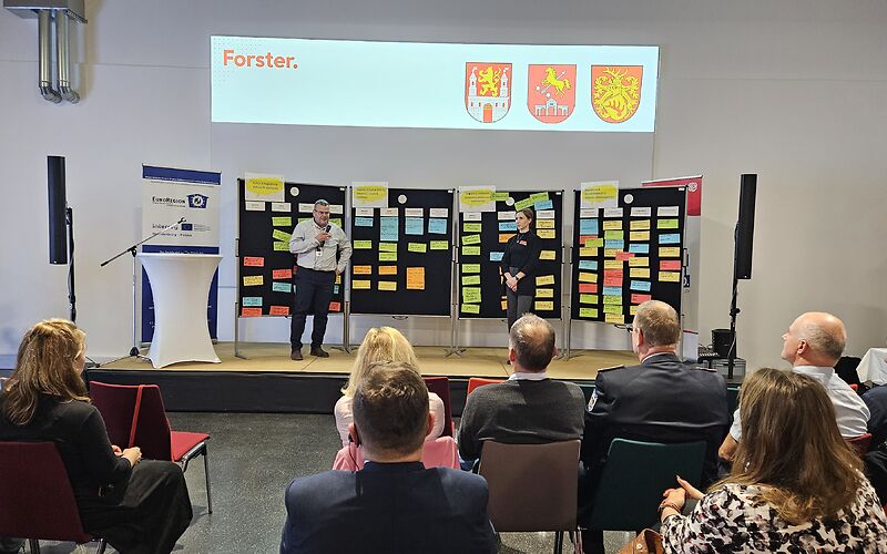 Auf der Bühne stehen ein Man und eine Frau, hinter ihnen sind 4 Pinnwände, an denen bunte beschriebene Zettel angepinnt sind. Vor der Bühne sitzen die Teilnehmenden in Stuhlreihen.