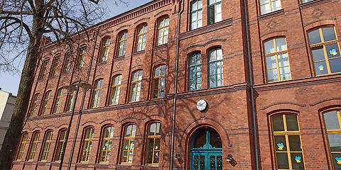Grundschule Nordstadt, roter Backsteinbau, 3 Etagen, Fensterfront mit gelben Fensterrahmnen rechts und links vom Eingang, Fenster über dem Eingang mit dunkelgrünen Fensterrahmen,Sicht auf den Eingangsbereich, eine große dunkelgrüne zweiflügelige verzierte Holtztür mit nach oben abgerundeten Oberlichtern