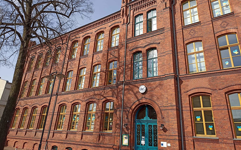 Grundschule Nordstadt, roter Backsteinbau, 3 Etagen, Fensterfront mit gelben Fensterrahmnen rechts und links vom Eingang, Fenster über dem Eingang mit dunkelgrünen Fensterrahmen,Sicht auf den Eingangsbereich, eine große dunkelgrüne zweiflügelige verzierte Holtztür mit nach oben abgerundeten Oberlichtern