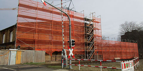 Zu sehen ist ein Anbau des Textilmuseums, welches durch ein Baugerüst verdeckt wird. Davor befindet sich ein Bahnübergang mit einer Lichtanzeige, Schranke und mit einem Andreaskreuz. An der Seite ist ein Teil der Badestraße zu sehen, welche entlang des Textilmuseums verläuft. Links am Textilmuseum steht eine Absperrung aus Holz.