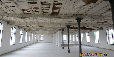 Blick in die leere Fabriketage. Alle nicht tragenden Wände sind zurück gebaut, einzig die Schienen der Rollschrankanlage des Archivs befinden sich auf dem Fußboden. An der Decke wurden die Holzwolle Leichtbauplatten zurückgebaut. Darunter sieht man die Lehmstaken in der Deckenkonstruktion.