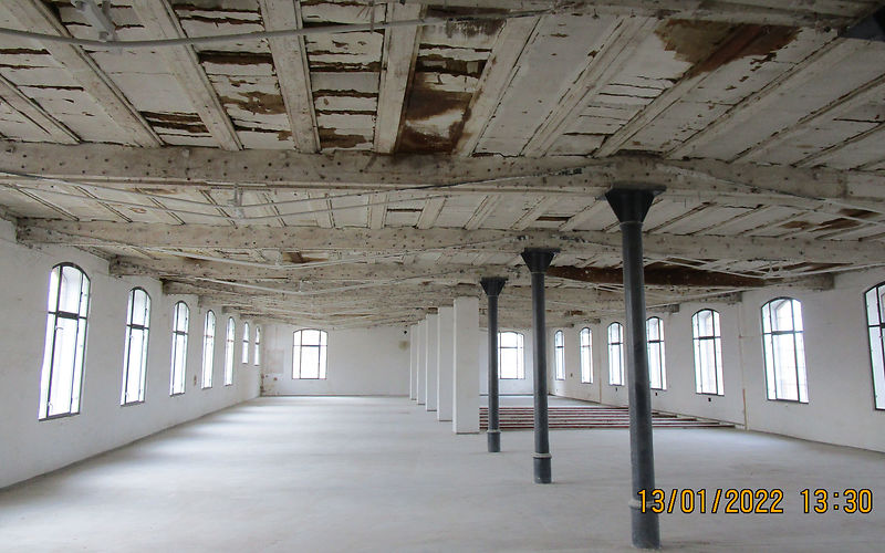 Blick in die leere Fabriketage. Alle nicht tragenden Wände sind zurück gebaut, einzig die Schienen der Rollschrankanlage des Archivs befinden sich auf dem Fußboden. An der Decke wurden die Holzwolle Leichtbauplatten zurückgebaut. Darunter sieht man die Lehmstaken in der Deckenkonstruktion.