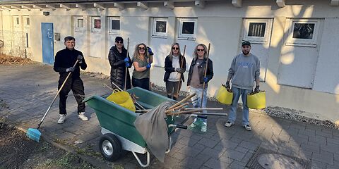 Das Team aus dem Hort Pfiffikus steht hinter einer grünen Schubkarre, die mit Arbeitsgeräten beladen ist. Im Hintergrund ist ein längliches einstöckiges flaches Gebäude. 