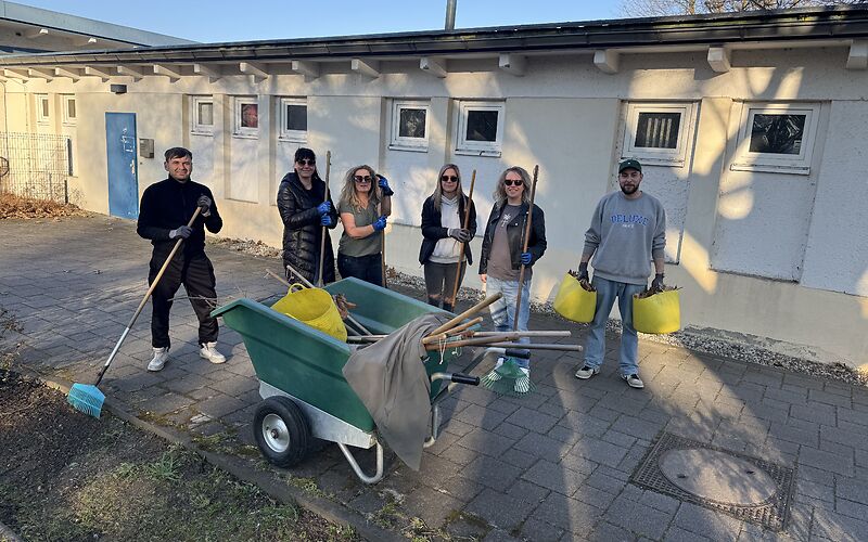 Das Team aus dem Hort Pfiffikus steht hinter einer grünen Schubkarre, die mit Arbeitsgeräten beladen ist. Im Hintergrund ist ein längliches einstöckiges flaches Gebäude. 