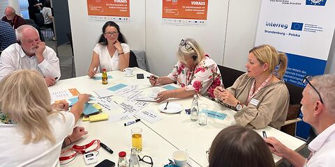 Frauen und Männer sitzen im Kreis am Tisch, vor ihnen bunte beschriebene Karten