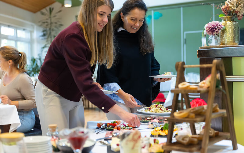 zwei Damen stehen vor einem reichgefüllten Frühstücksbuffet und tragen sich einzelnen Häppchen auf den Tellern auf