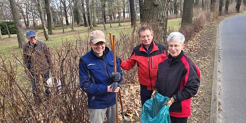 4 erwachsene Personenstehen mit Müllsack und Harken an einem Seitenstreifen einer Straße, auf dem kahle Büsche stehen. Auf dem Boden liegen Blätter.