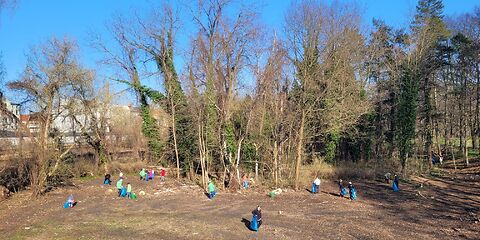 Mehrere Erwachsene und Kinder Sammeln vor einer kleinen Wald mit blauen Müllsecken Müll ein. 
