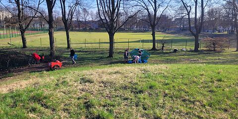 Auf der Wiese vor einem Sportplatz sind mehrere Personen mit Bollerwagen, die Müll sammeln.