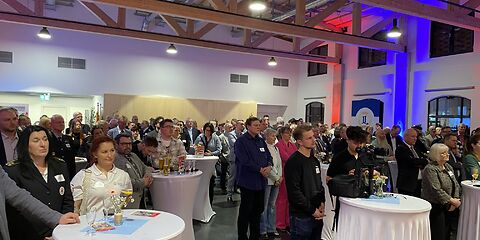 Blick von vorn in den gefüllten Saal, die Gäste stehen in Gruppen an Stehtischen mit weißen Tischdecken.